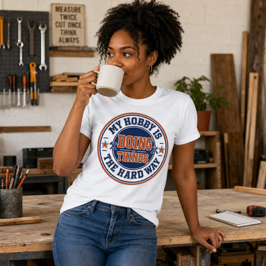 A woman in a workshop setting wearing a white graphic t-shirt with the text 'MY HOBBY IS DOING THINGS THE HARD WAY' in blue and orange, paired with blue jeans.